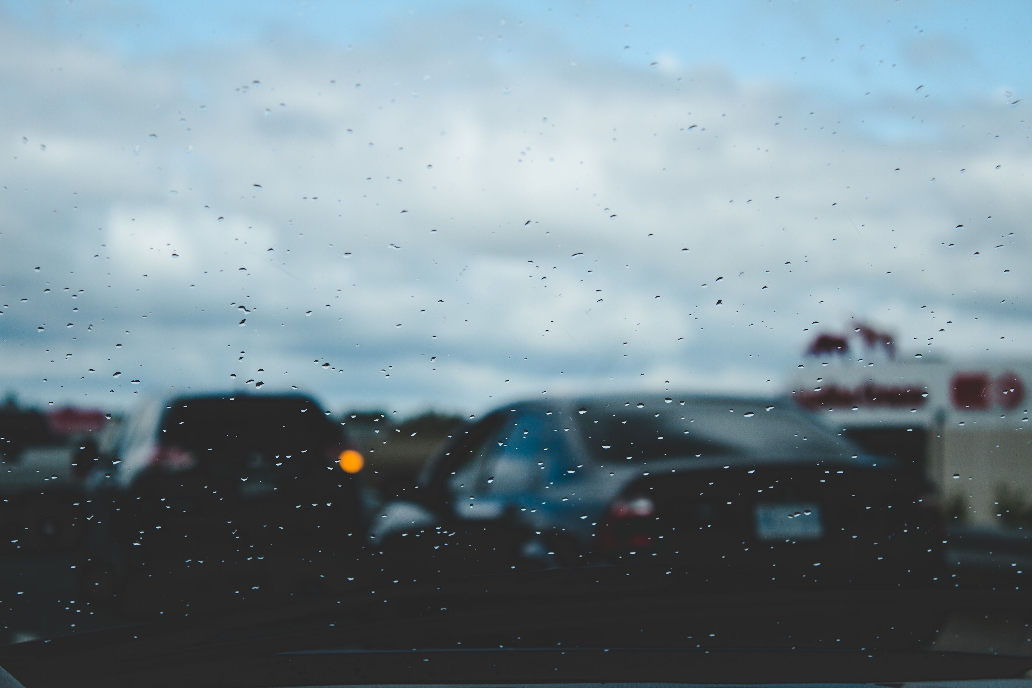 A moody photo showing raindrops on a car window, creating a soft focus effect on the blurred traffic outside. The overcast sky and wet conditions add a sense of melancholy and reflection, capturing a quiet moment in the hustle of everyday life.