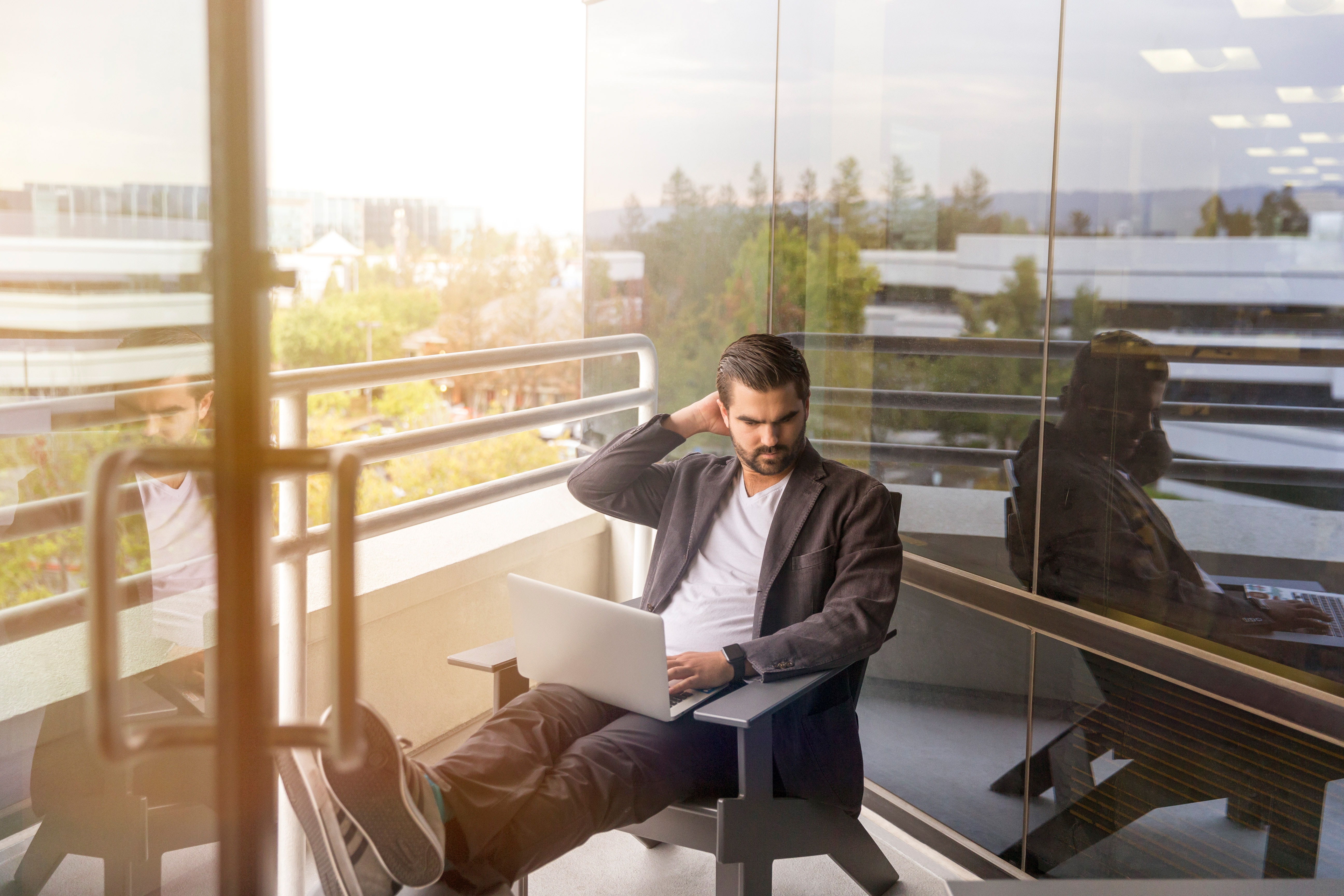 Man on a computer and sitting on a balcony with sunlight behind.